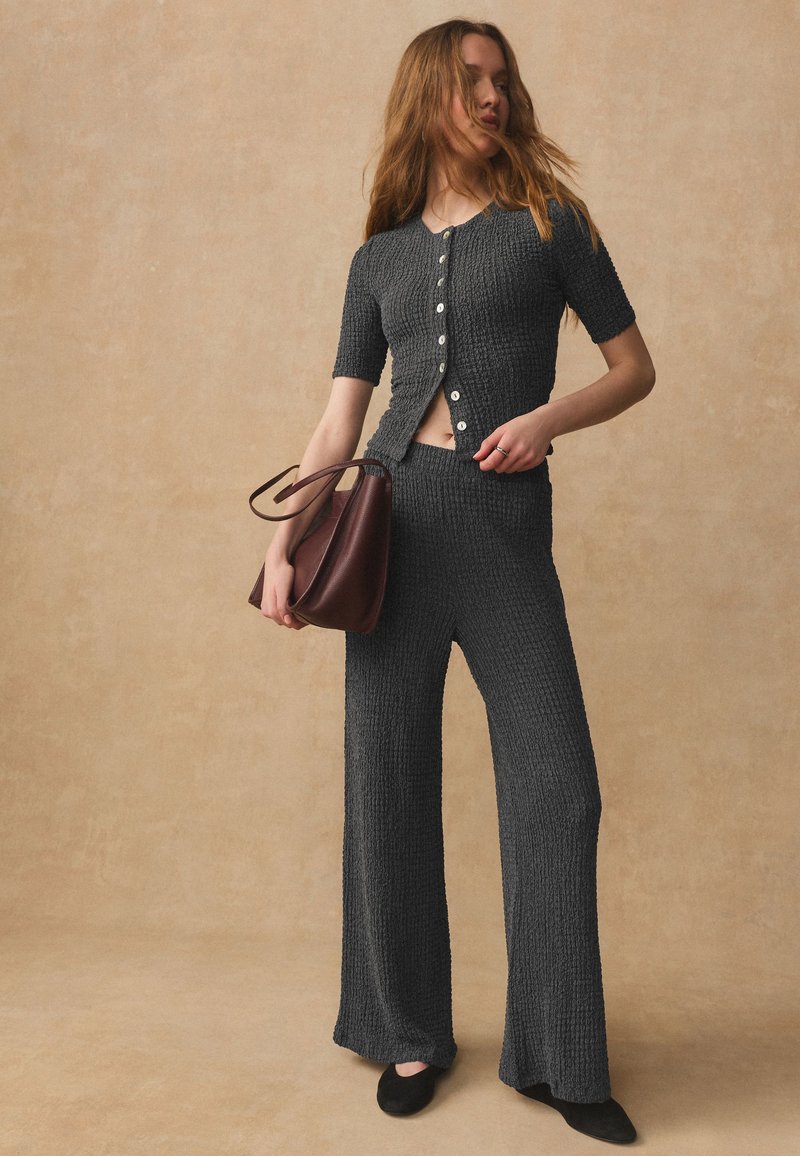 Femme portant un haut boutonné à manches courtes gris foncé texturé et un pantalon large assorti, tenant un sac à main marron, debout devant un fond beige.
