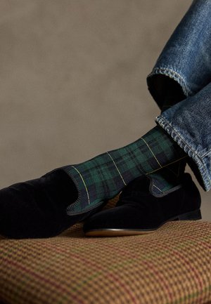 Svarte fløyelsloafers hvilende på en rutete polstring, kombinert med grønne og blå rutete sokker med gule linjer. Denimjeans er synlige.