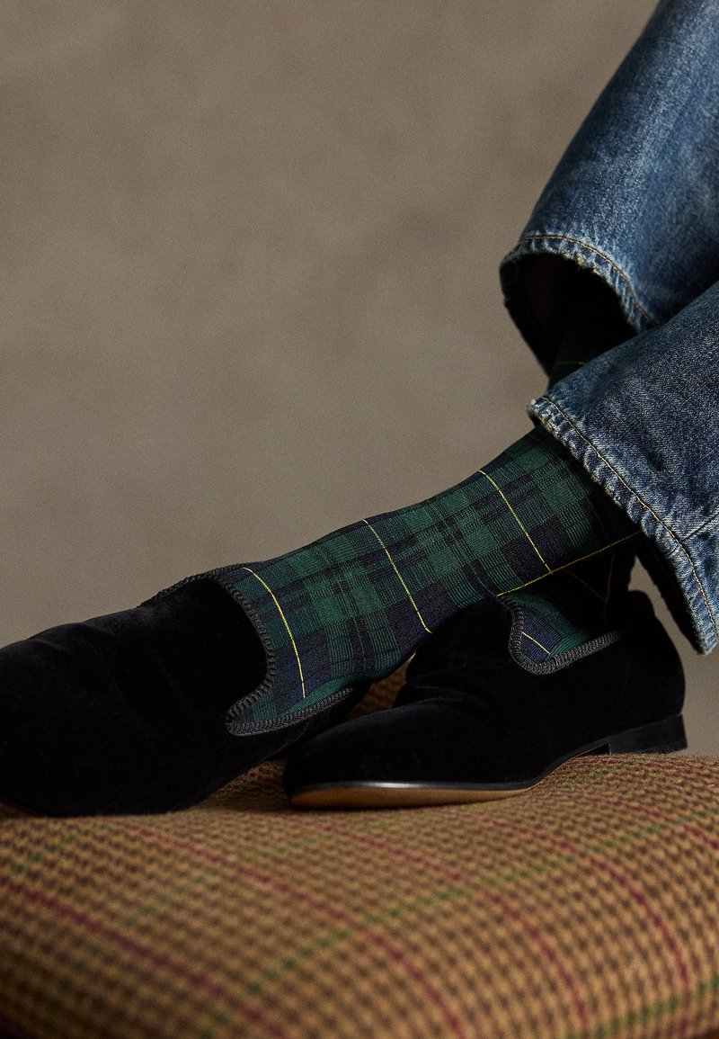 Svarte fløyelsloafers hvilende på en rutete polstring, kombinert med grønne og blå rutete sokker med gule linjer. Denimjeans er synlige.