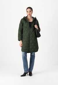 Olivgrüner gesteppter langer Mantel mit Kapuze, ausgestattet mit einem Reißverschluss auf der Vorderseite, seitlichen Taschen und einer lockeren Passform. Kombiniert mit blauen Jeans und schwarzen High Heels.