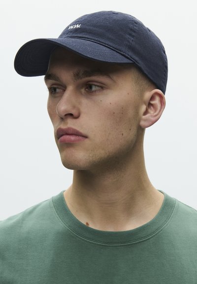 Junger Mann mit dunklem blauen Cap und grünem Rundhalsshirt, schaut nach links vor einem schlichten hellen Hintergrund.