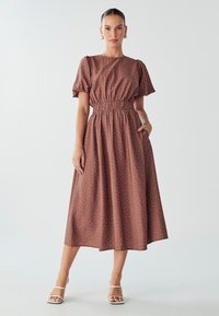 Smeđa midi haljina s točkicama, nabranim rukavima, strukom i bočnim đepovima. Izrađena od laganog materijala, u kombinaciji s bijelim sandalama na petu.