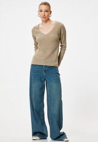 Beige gerippter Langarmpullover mit V-Ausschnitt, kombiniert mit weiten blauen Jeans und weißen Sneakers. Glattes Material und lockere Passform.