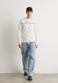Weißes Langarmhemd mit schwarzem horizontalen Streifen und Text, hellblaue, locker sitzende Jeans, schwarze Cap und schwarze Schuhe. Einfaches, lässiges Outfit.