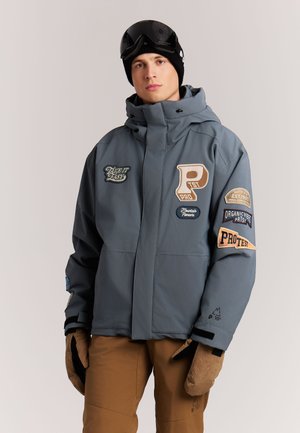 Jonge man draagt een grijze winterjas met capuchon en patches, een bruine broek, beige handschoenen, een zwarte muts en skibril op het voorhoofd.