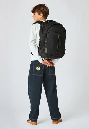 Junge trägt weißen Pullover und dunkle Hose mit Smiley-Aufnäher, trägt einen großen schwarzen Rucksack und steht vor einem einfarbigen Hintergrund.