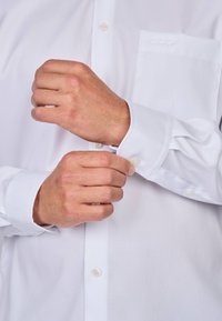 Chemise blanche à manches longues, dotée d'une poche poitrine unique et de boutons transparents. Texture lisse, poignets bien ajustés.