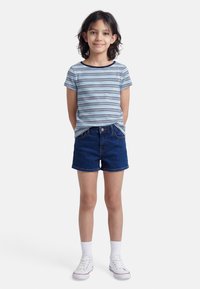 Νεαρή κοπέλα που στέκεται με τα χέρια πίσω από την πλάτη, φορώντας μπλε ριγέ μπλουζάκι, τζιν σορτς, λευκές κάλτσες και λευκά αθλητικά παπούτσια.