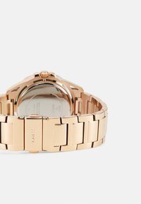 Orologio in oro rosa con bracciale in metallo a maglie, cassa rotonda e finitura lucida. Presenta un logo visibile e dettagli in metallo testurizzati.