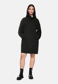 Schwarzes, oversized Kleid mit hohem Kragen und gerippten Akzenten. Verfügt über lange Ärmel und seitliche Taschen, getragen mit klobigen schwarzen Stiefeln.