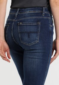 Jeans de mezclilla azul oscuro con corte ajustado. Incluyen una cinturilla estándar, bolsillos traseros y detalles de costura sutiles.