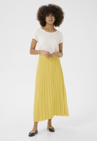 Gonna midi plissettata gialla, abbinata a una t-shirt bianca a scollo rotondo. La modella indossa delle ballerine nere. Tessuto morbido, design fluido.