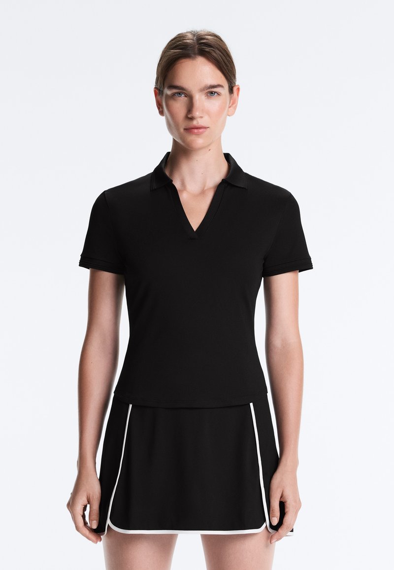 Femme aux cheveux tirés en arrière, portant un polo noir à manches courtes et une jupe noire avec un liseré blanc, debout sur un fond uni.