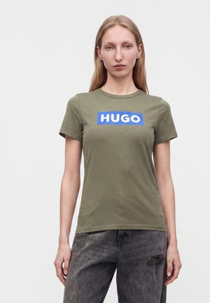 Млада жена с дълга руса коса, облечена в маслинено зелена тениска с "HUGO" в бяло върху синьо и избелели черни скъсани дънки.