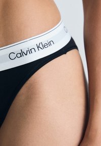 Crne tange s bijelim elastičnim pojasom na kojem se nalazi "Calvin Klein" logo. Glatka tekstura, niska linija struka, minimalistički stil.