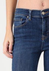 Dark blue denim jeans with a fitted cut, front pockets, and visible yellow stitching along seams; waistband with metal button closure.