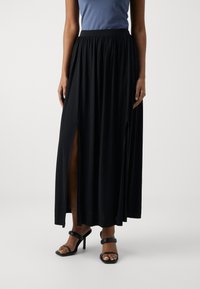 Žena v černé maxi sukni se dvěma předními rozparky, doplněná černými sandály na vysokém podpatku a modrým topem bez rukávů.