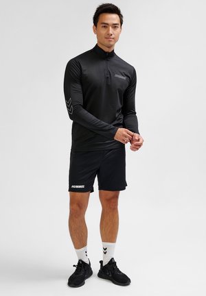 Zwarte lange mouwen shirt met een kwart rits en zij-chevron detail, gecombineerd met zwarte shorts en zwarte sport schoenen.