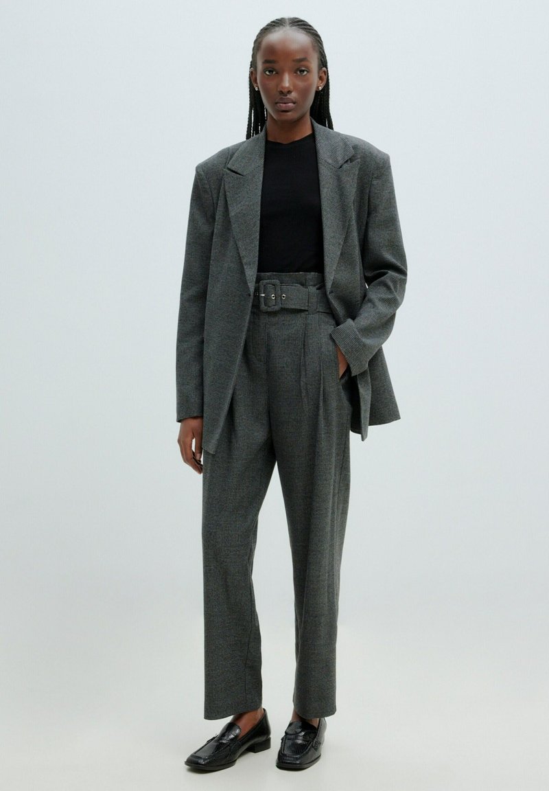 Grau kariertes Blazer mit breiten Revers, hochgerafften Hose mit Gürtel und schwarzen Slippern. Oben ist ein figurbetontes schwarzes Hemd.