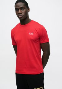 Piros rövid ujjú póló kerek nyakkivágással és kis fehér EA7 logóval a bal mellrészen. Sima anyagú, testhezálló fazon.