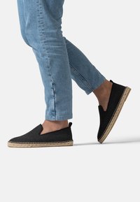 Černé slip-on boty s texturovaným textilním svrškem a tkanou juteovou podešví. Světle modré džíny s kratší délkou na kotníky.