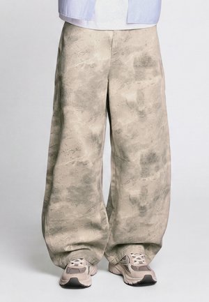 Lockere beige Hose mit einem strukturierten Muster, getragen mit beige-weißen Sportsneakern auf schlichten Hintergrund.