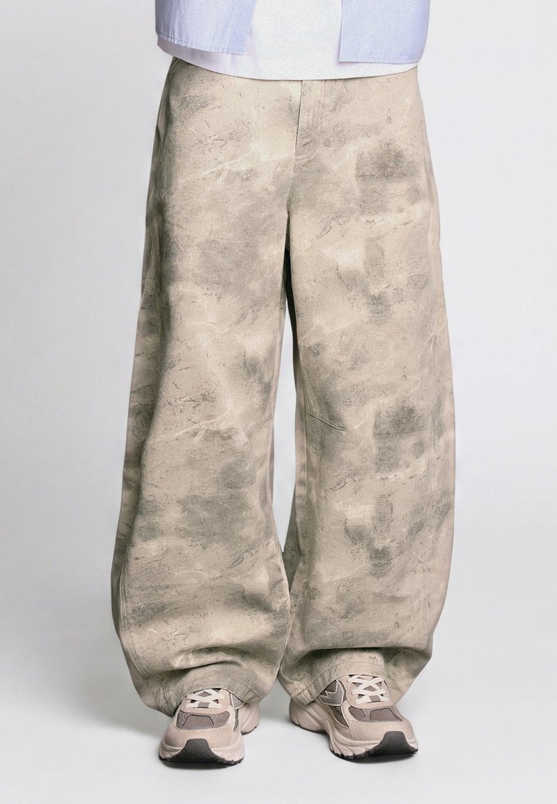 Pantalon beige large avec un motif texturé porté avec des baskets de sport beiges et blanches sur un fond uni.