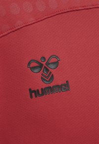 Czarne logo Hummel z motywem trzmiela nadrukowane na czerwonej teksturowanej tkaninie z przeszytym szwem i kropkowanym wzorem w pobliżu krawędzi.