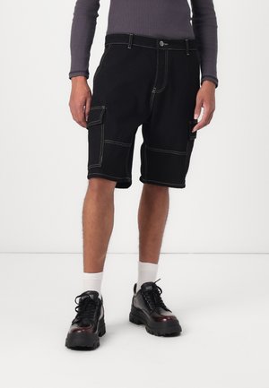 Shorts cargo noirs avec des coutures contrastées, dotés de multiples poches et d'une coupe décontractée, associés à des chaussures noires robustes et des chaussettes blanches.