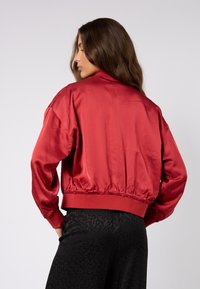 Czerwona satynowa kurtka bomberka z elastycznym pasem i mankietami, o gładkiej fakturze i swobodnym kroju. Noszona na spodniach w czarny nadruk w panterkę.
