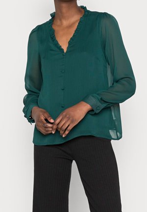 Vrouw draagt een donkergroene blouse met een gerimpelde kraag en stof bedekte knopen, gecombineerd met zwarte geribde broek, handen zachtjes gevouwen.