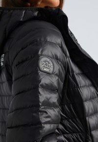 Svart nedjacka med kviltad design och puffade sektioner, dragkedjestängning och en rund logotyp-patch på vänster ärm.