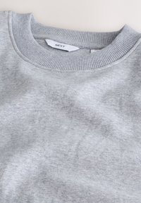 Svijetlosiva majica dugih rukava s okruglim izrezom, rebrastim ovratnikom i bijelom etiketom s oznakom "NEXT" unutar izreza.