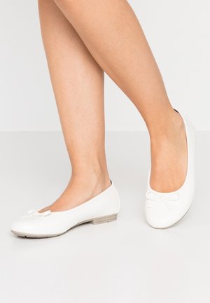 Femme portant des ballerines blanches avec de petits nœuds, debout sur un sol gris clair devant un fond blanc.