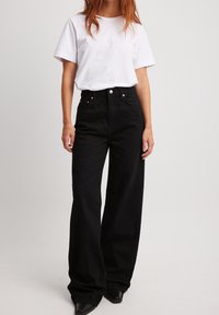 Svarta högmidjade wide-leg jeans i denim, med framfickor, metallknappstängning och en slät textur. Bärs med en enkel vit t-shirt.