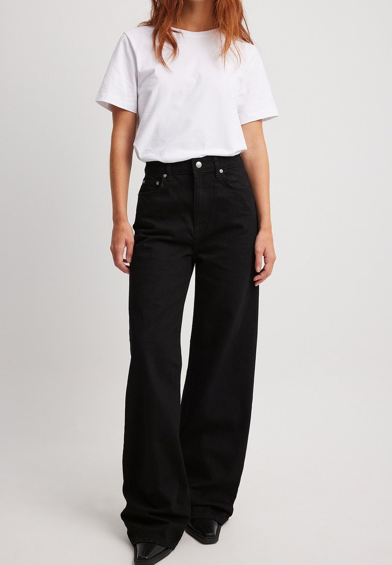 Svarta högmidjade wide-leg jeans i denim, med framfickor, metallknappstängning och en slät textur. Bärs med en enkel vit t-shirt.