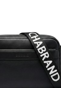 Sac bandoulière en cuir noir avec une finition texturée, doté d'une sangle proéminente avec un logo blanc et d'une fermeture zippée.