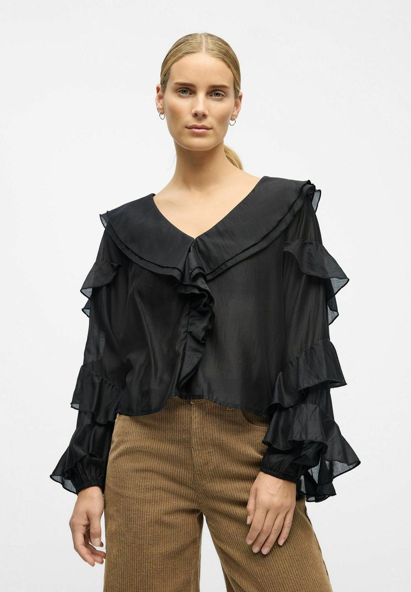 Femme aux cheveux blonds portant un chemisier noir à volants et un pantalon en velours côtelé marron, debout devant un fond clair et uni.