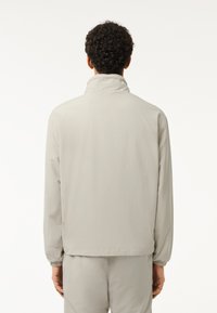 Hellgraue Jacke aus leichtem Stoff, mit Stehkragen, langen Ärmeln, elastischen Bündchen und einer schlichten, glatten Textur.