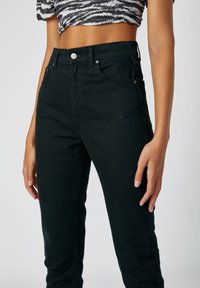 Černé džíny s vysokým pasem vyrobené z denimu. Mezi charakteristické prvky patří tradiční zapínání na knoflík a přední kapsy s jemným švem.