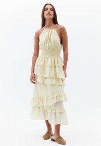 Záhybovitá, bezramínková žlutá šaty s roubeným pasem a volánkovými vrstvami. Vyrobené z lehkého materiálu, doplněné pletenými nazouvacími botami.