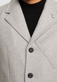 Abrigo de mezcla de lana gris con solapa muesca, dos botones negros y un bolsillo en el pecho que presenta un sutil alfiler. Suéter negro debajo.