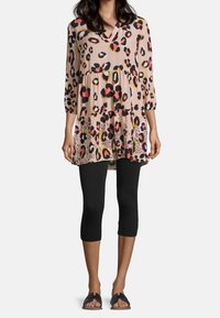 Túnica de manga larga y escote en V en una suave y ligera tela con un fondo beige, que presenta un estampado de leopardo colorido, combinada con leggins negros y sandalias.