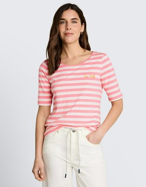Kobieta ubrana w różowo-biały, poziomo paskowany t-shirt z krótkim rękawem, w połączeniu z jasno beżowymi spodniami z sznurkiem w pasie, stoi z jedną ręką w kieszeni.