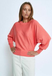 Korallfärgad oversized tröja i mjukt material, med rund halsringning och långa, vida ärmar. Matchas med ljusa byxor.