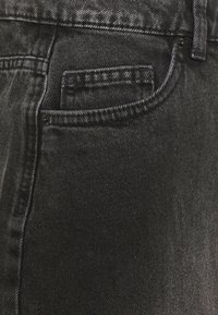 Gros plan sur une poche de jean en denim noir avec poche à monnaie, une passants de ceinture visible et des rivets sur un tissu texturé.