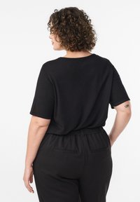 Melns īss piedurkņu tops ar vaļēju piegriezumu, apaļu izgriezumu un mīkstu tekstūru, kombinēts ar melnām elastīgām biksēm ar kabatām.