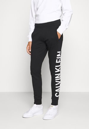 Osoba nosząca czarne spodnie dresowe Calvin Klein z dużym białym logo na nodze, białą bluzkę z długim rękawem i białe trampki, stojąca z jedną ręką w kieszeni.