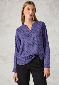 Blusa lunga viola con un motivo a diamante, caratterizzata da un scollo a V e una vestibilità morbida, abbinata a pantaloni neri. Tessuto dalla superficie liscia.