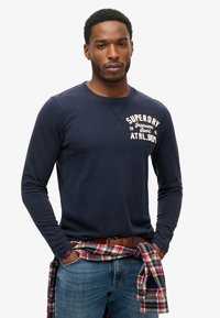 Navy long-sleeve shirt with a crew neck and front graphic text. Worn with light blue jeans and a red plaid flannel tied around the waist.